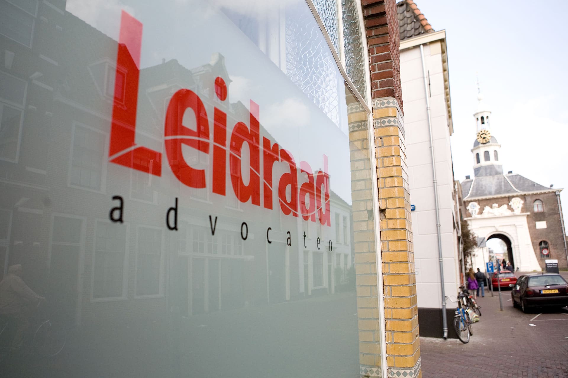 De naam Leidraad advocaten op het raam van het kantoor, met een oude stadspoort in Leiden op de achtergrond